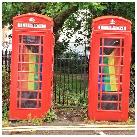 Two red BT telephone boxes that have the progress pride flag hanging up inside.