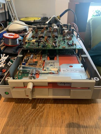 An opened 1541 floppy drive, showing an orange foppy disk inserted, the main PCB with a little perfboard bodge and many electronic components.