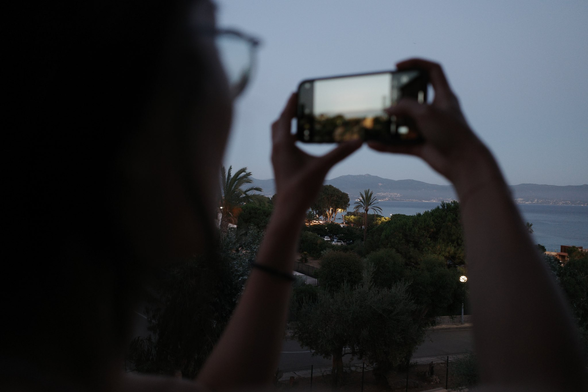 A night time view of the sea with someone taking a photo of it on their phone in the foreground