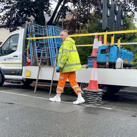 contractor stood next to a flat bed van while wearing hi-vis yellow jacket, orange trousers tucked into pristine white socks and trainers