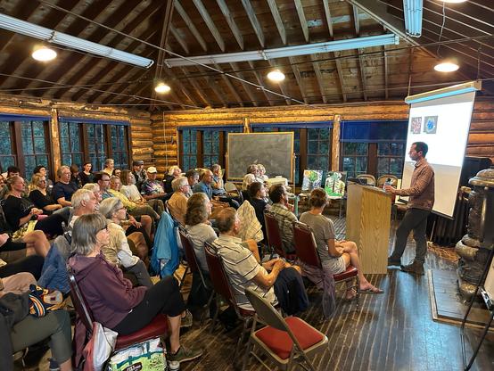 A crowd sitting closely together listens to a speaker present slides in the Megaron building at CU Boulder's Mountain Research Station. This old building has beautiful exposed log walls as well as a pyramid-shaped wooden ceiling and dark wooden floor.