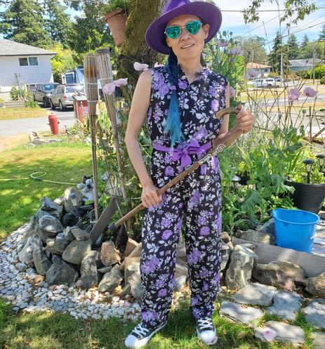 Photo by my Mom.
Wearing my purple floral jumpsuit, rainbow black & white sneakers, heart shaped sunglasses, purple hat & holding my yew wood cane, in our front gardens.
