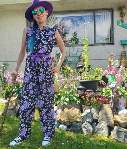 Photo by my Mom.
Wearing my purple floral jumpsuit, rainbow black & white sneakers, heart shaped sunglasses, purple hat & holding my yew wood cane, in our front gardens.