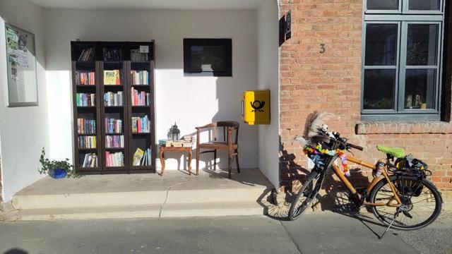 Ehemalige Haltestelle mit einem Bücherschrank, einem kleinen Tisch und einem Stuhl, sowie einem gelben Postbriefkasten. Daneben steht ein Fahrrad mit bunt geschmücktem Lenkerkorb