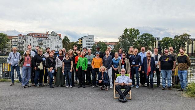 Eine gemischte Gruppe steht vor einem Fluss und Städte Hintergrund 

Vorne sitzt ein älterer Heer auf einem Sonnenstuhl