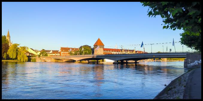 Blick auf die Stadt vom anderen Ufer aus. Ein Turm erhebt sich im Zentrum und rechts daneben führt eine Brücke über den Fluss.