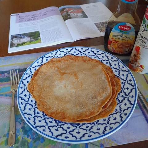 A plate with thee thin pancakes on top of each other. To the right are some toppings and behind the plate  a magazine is flipped open.
