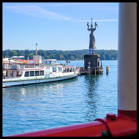 Blick auf eine Anlegestelle. Ein Schiff hat angelegt und rechts daneben ist eine Statue zu sehen. Im Hintergrund erheben sich Berge.