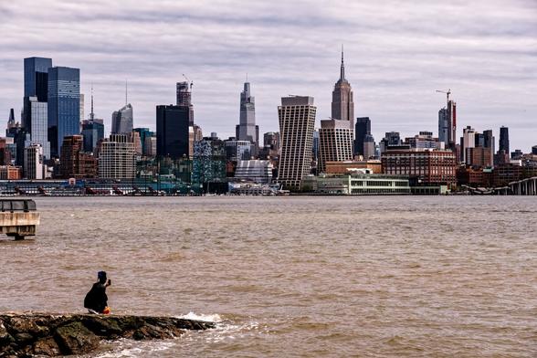 A color photograph taken from the Hoboken waterfront, looking across the Hudson River at Manhattan. On the left, a woman is crouching on a rocky outcrop in the river on the near shore, taking a photo or video of the Manhattan riverfront. The Hudson has a muddy tint this close to its western bank. The Empire State Building sits in the background just to the right of center among the collection of buildings, and the light green paint of the Pier 57 building stands out along the shoreline.  On the right edge of the frame,  a portion of City Island is visible.