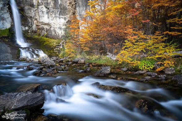 Long exposure photograph of a waterfall in the background and the outflow river in the foreground. On the right side is a forest with bright orange fall foliage