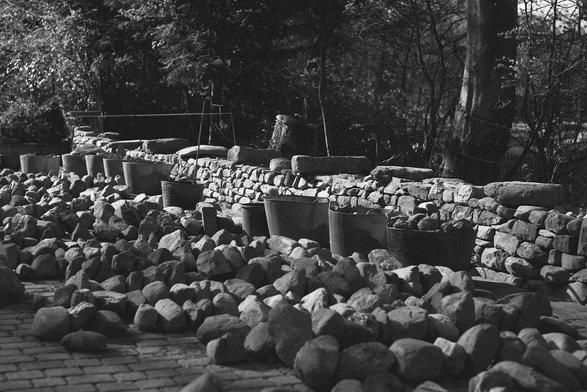 A black and white photograph of a partially built drystone wall. There are stones on the ground in lines and plastic buckets and a half built drystone wall behind them.