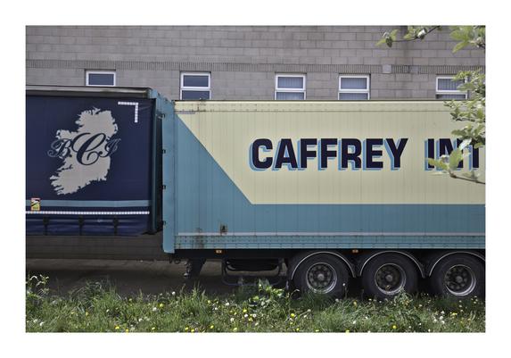 A close-up view of two adjacent truck trailers parked side by side. The trailer on the left is dark blue with an image of Ireland and the initials "BCJ." The trailer on the right is light blue and yellow with the text "CAFFREY INT" partially visible. The trailers are parked against a gray brick building with small windows near the roofline. In the foreground, there is a patch of grass and wildflowers.
