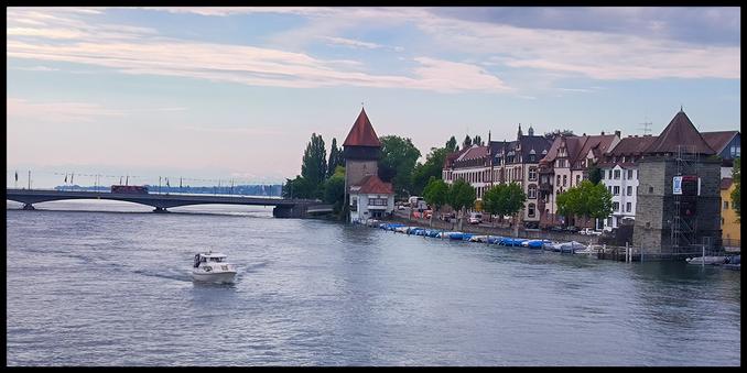 Blick auf ein paar Gebäude. Links davon führt eine Brücke über den Fluss. Auf dem Fluss fährt ein Boot. Der Himmel ist leicht bewölkt.