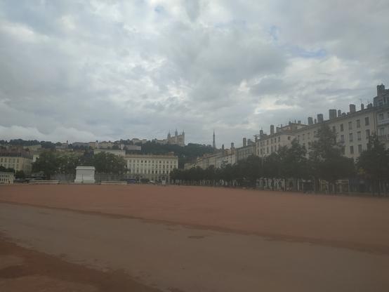 Place Bellecour, we see the redish ground, statue of Louis dont-remember-the-number, building, and my objective : the basilique of Fourvière and the Radio Tower, All this under a grey sky