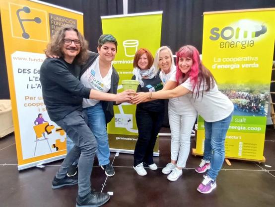 Cinc persones uneixen les mans. De fons, es veuen cartells de Coop57, Germinador Social i Som Energia.