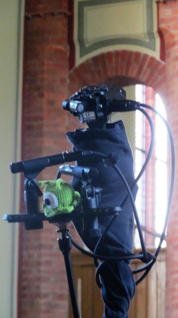 Auf einem mit schwarzem Stoff umwickelten Ständer ist ein Fieldrecorder mit XY-Kapseln befestigt. Davor wird eine Stereoschiene von einem Arm gehalten. Auf der Stereoschiene sind zwei Kondensatormikrophone in ORTF-Stellung angebracht. In der Mitte dazwischen sitzt eine gehäkelte Gehirnschnecke. Sie ist klein, kugelig und grün, hat ein Auge und zwei Fühler.
Im Hintergrund ist gemauertes Gewölbe von der Halle zu sehen.