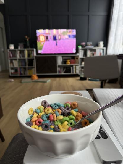 A white bowl full of froot loops sitting on a small white table. In the background you can see a tv with the video game Disney Dreamlight Valley loaded on the screen