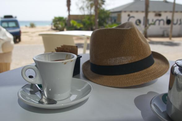 Hat and coffee on a table, the beach in the far, blurry background behind a building and a car