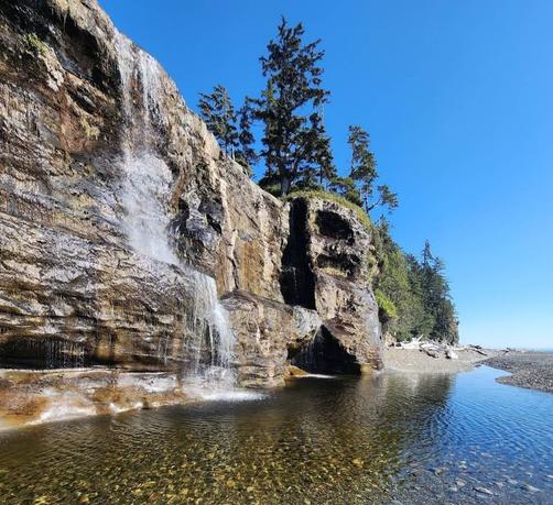 A waterfall runs down large rock cliff, pooling below with oceanfront beach beside it.
Trees are atop the cliff & forest beside beach is visible in far background.