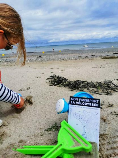 Enfant sur une plage de sable, pelle et râteau verts au premier plan, passeport Vélodyssée