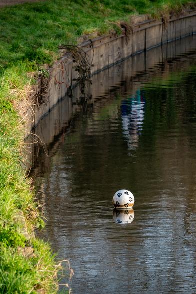 A football floats on the water next to a canal. A hazy reflection of a person is visible beyond it.