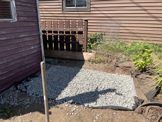 an area about 7' x 4' is covered in 1 inch Riverrock. It is relatively flat. There is a fading purple shed to the left. A temper stands in the front of the picture. Behind is a brown fence and brown siding of a garage.