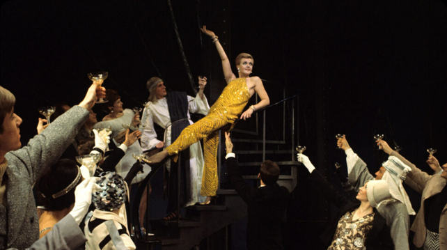 A photo from the original Broadway production of Mame, its opening number of sorts, "It's Today". Against a black background, Auntie Mame is posing center in a yellow pants outfit (bare arms), with the cast raising their glasses towards her.