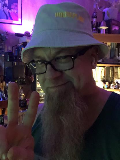 Me in the studio, darkly shadowed against a brightly lit background. I am flashing a peace sign with my fingers and have on a light blue bucket hat with gold lettering that reads: HOUSEPLANT