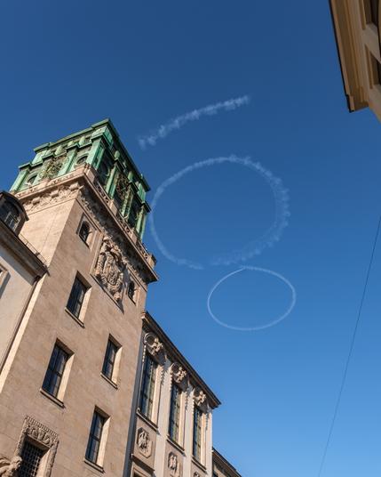 The old clock tower of the TU Munich with a crystal clear sky above it. Our yellow acrobatics plane "Schlacro" is flying above, painting a large "100" into the sky with white smoke.