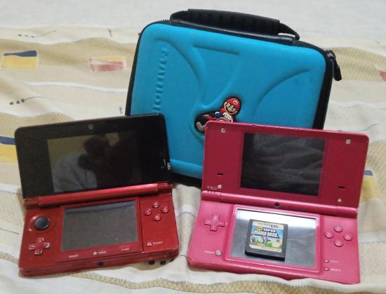 A photo of my DS collection, a red OG Nintendo 3DS and a pink Nintendo DSi, a Mario Kart 7 carrying case (which came with the 3DS) is behind the consoles and a New Super Mario Bros. DS cartridge is on the DSi's bottom screen.