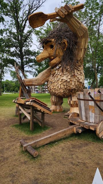 A very large wooden troll stands in a park. His right hand is stuck in a "portal."
A large cart and bucket in foreground. Trees in background.