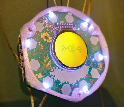 An EMF Tildagon badge with a top bumper fitted, the bumper is a translucent colourless plastic with glittery flecks in it, the LEDs are alternately shining white and glowing through the bumper.