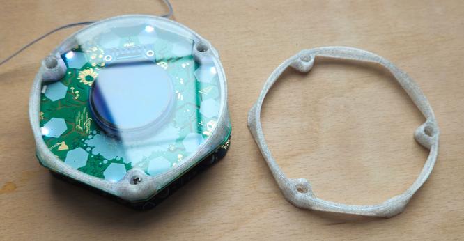On the left an EMF Tildagon badge with a top bumper fitted, the bumper is a translucent colourless plastic with glittery flecks in it, the LEDs are alternately shining white and glowing through the bumper. 

On the right a print of the bumper on its own.