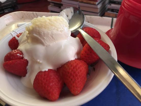A generously filled bowl full of big red strawberries, extra thick cream and Cornish clotted cream on top, plus a large soup spoon to eat it all with, and a red drink tumbler beside the bowl