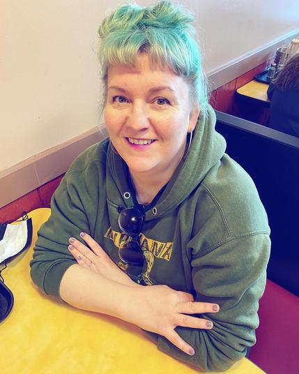 Agni Hogaboom, a nonbinary artist in Washington state, smiles at the camera. They have blue-green hair, wear a green hoodie, and have arms folded on a yellow table.
