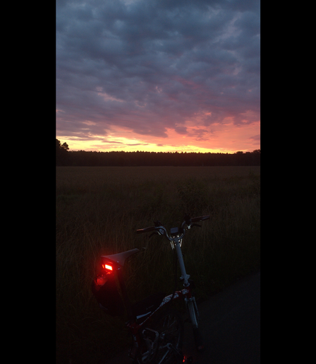 Ein halb zusammengefaltetes Brompton-Faltrad steht vor eoner noch dunklen Weide. Ein Rücklicht am Sattel leuchtet. Am Horizont der sich anbahnende Sonnenaufgang von hellgelb über pampelmußefarben bis hin zu blaugrauen Wolken.