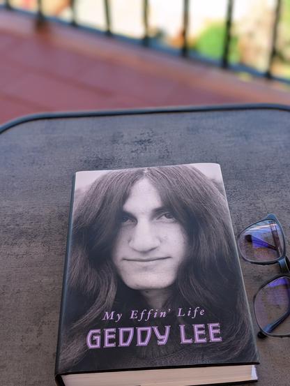 The image shows a book titled "My Effin' Life" by Geddy Lee. The cover features a black-and-white photograph of a young Geddy Lee with long hair, looking directly at the camera. The title and author's name are written in purple font. The book is placed on a gray surface, with a pair of eyeglasses next to it. In the background, there is an out-of-focus railing and some greenery, suggesting an outdoor setting.