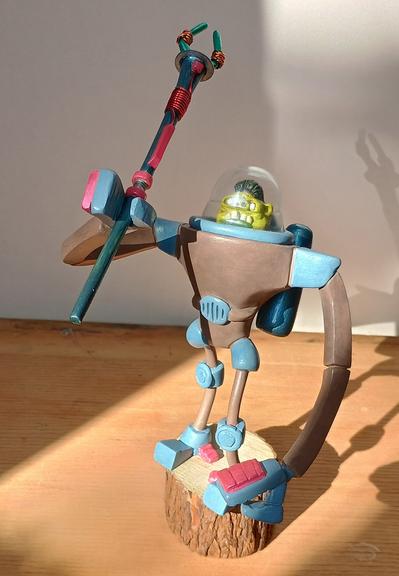 A figurine with a robotic body and green organic head. It's got a transparent helmet and is holding aloft a two pronged spear.