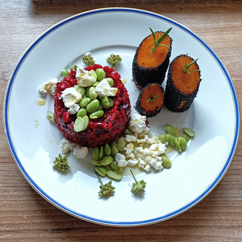 eine rund angerichtete Portion des Risottos, bestreut mit Edamame und Feta. Auf dem Teller stehen noch wie drei kleine Türme Karottenstücke, die außen mit Mohn ummantelt sind.