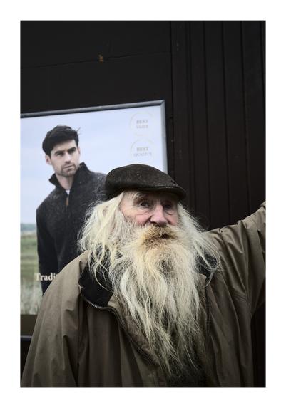 An elderly man with a long, flowing white beard and a flat cap poses for the camera. He is wearing a brown coat and stands in front of a dark wall. Behind him, a poster featuring a young man with dark hair and a beard advertises clothing with the text "BEST VALUE" and "BEST QUALITY." The contrast between the two men—the elderly gentleman and the younger model—creates an interesting visual dynamic.
