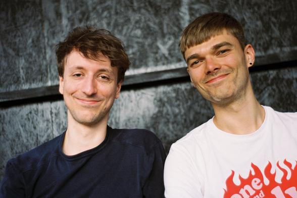 A photo of two guys. One has darker and longer hair and a dark t-shirt, while the second has a brighter and shorter hair and a white t-shirt. They look straight into the camera and smile.
