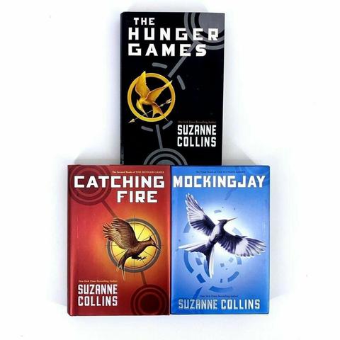 Three books with a light gray background.

The first book has a black cover, and has a gold pin shaped like a bird in a circle holding an arrow. The title reads: The Hunger Games. The author is Suzanne Collins.

The second book has a red cover, and a gold circle in the center, with a brown-gold bird flying. The title reads: Catching Fire. The author is Suzanne Collins.

The third book has a blue cover, with a black and light gray bird flying against a broken circle. The title reads: Mockingjay. The author is Suzanne Collins.