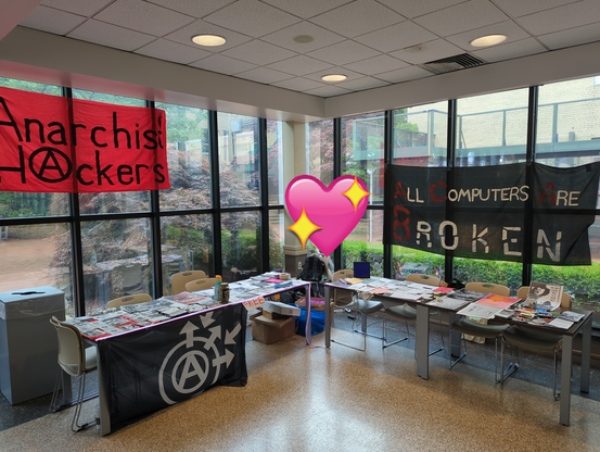Anarchist Hacker Village at HOPE XV. There are tables with various zines, stickers and posters for free, with banners reading "Anarchist Hackers" and "All Computers Are Broken"