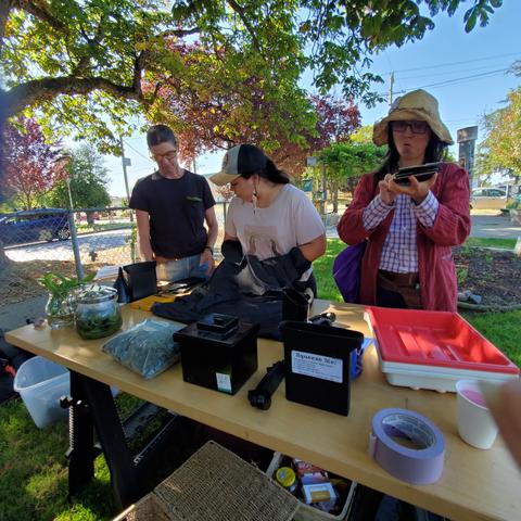 Suzi, one of the artist instructors, helping a workshop attendee learn how to get silver plate film ready for shooting. Attendee has both hands inside a large black bag. Another attendee is looking at a film box.