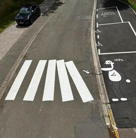 Streetart. A crosswalk has been painted on an asphalt road and a cartoon character next to it. It is a simply drawn little man with a protruding lower jaw, one large and one small eye, thin arms and legs. He is standing at the side of the road with his mouth open and has just tapped the crosswalk (also drawn) with one finger, playing dominoes. The result is two tilted stripes that plop onto the three straight stripes. Next to them is the word "Oops". A great mixture of street art in the truest sense of the word and installation.