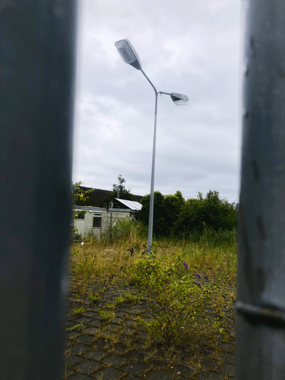 Seen between two metal poles, an abandoned parking lot overrun with weeds. In the center of the frame is a tall pole with two large lights on it. In the background is a cloudy grey sky.