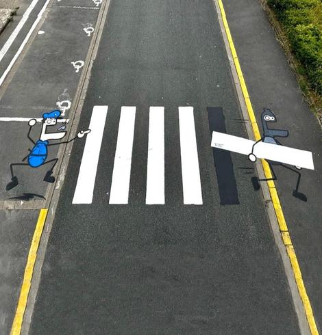 Streetart. A crosswalk with two funny cartoon figures has been painted on an asphalt road. They are two simply drawn little men with protruding lower jaws, one large and one small eye, thin arms and legs. On the right-hand side, an excited policeman in a blue uniform is jumping around and shouting after a black-masked robber on the left, who is in the process of carrying away a strip of the crosswalk. 
Info: The artist Toc Toc has designed several walls and crosswalks for the "Les Petits Bonheurs" festival. All unmistakably grandiose.