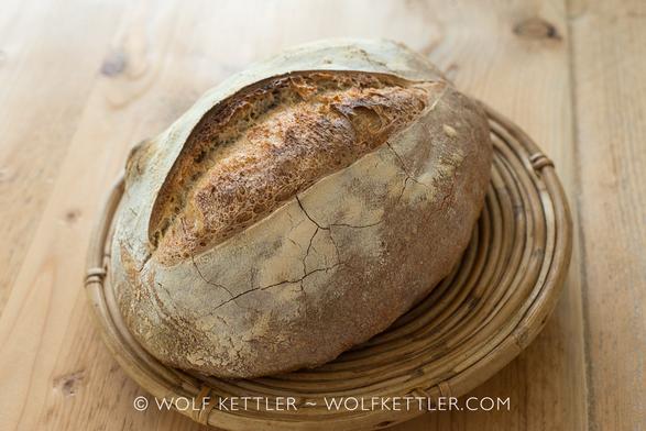 The photograph shows a scored loaf of crusty bread displayed on a rustic wicker plate. The plate itself sits on a wooden tabletop.