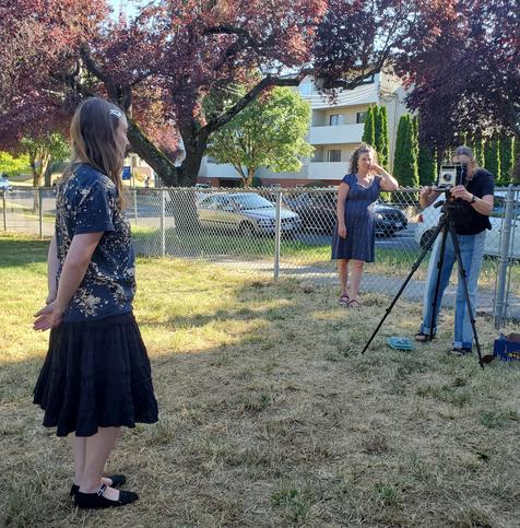 Graflex Crown Graphic large format camera, mounted on tripod, Suzi, is getting ready to take a portrait of an attendee, who is standing on the left. Another attendee is leaning against a fence, behind Suzi, to the right.