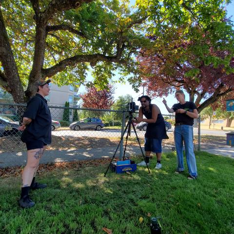Graflex Crown Graphic large format camera, mounted on tripod. Joshua is getting ready to focus & take a portrait of an attendee, standing to the left. Suzi, is beside Joshua, talking & pointing.